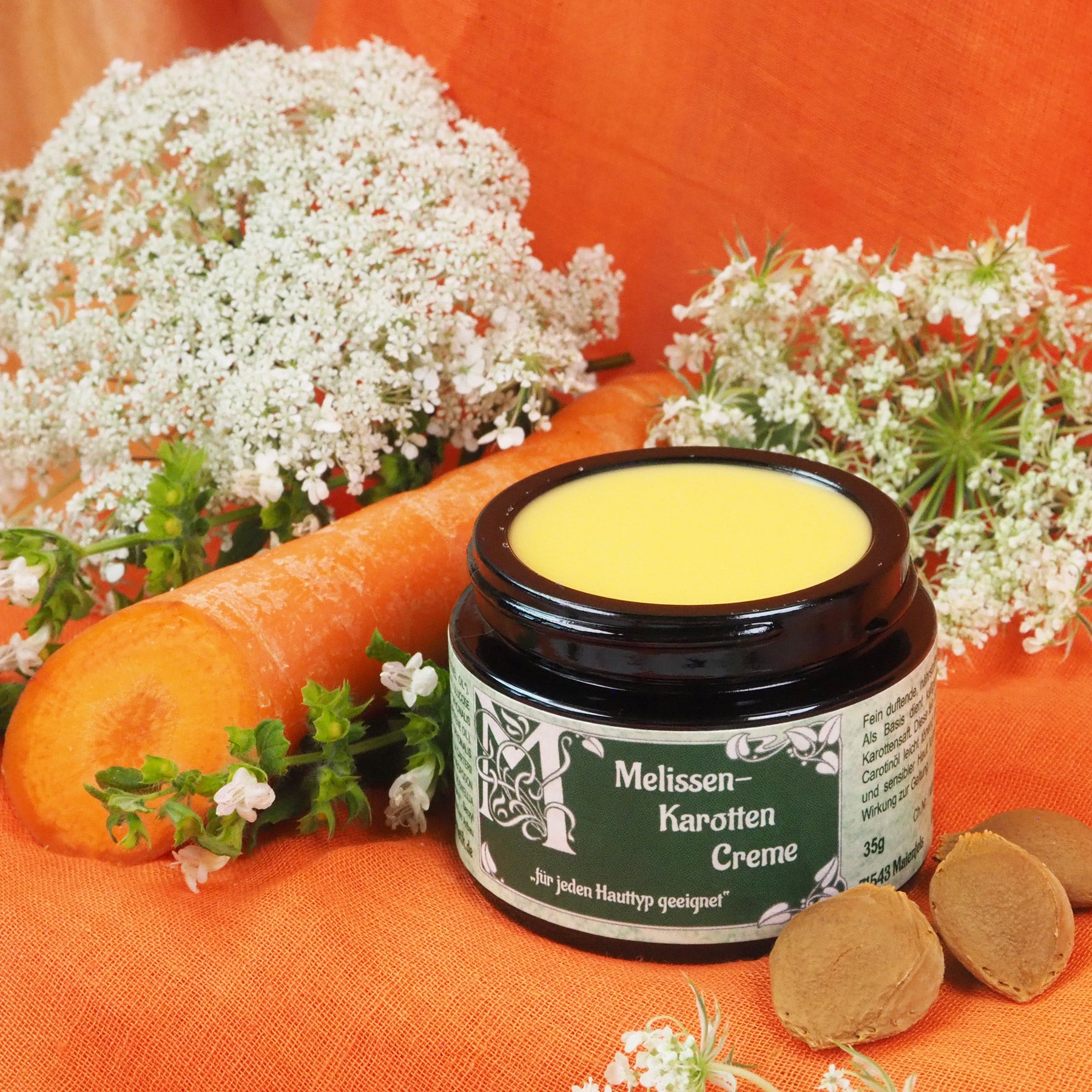 Geöffneter Melissen-Karotten Creme Tiegel 35ml. Daneben liegt eine Karotte.